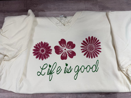 grit city rebel clothing unisex t-shirt in cream with the phrase life is good in green glitter vinyl and three flowers in pink glitter vinyl.  Available in sizes small through 3x. 