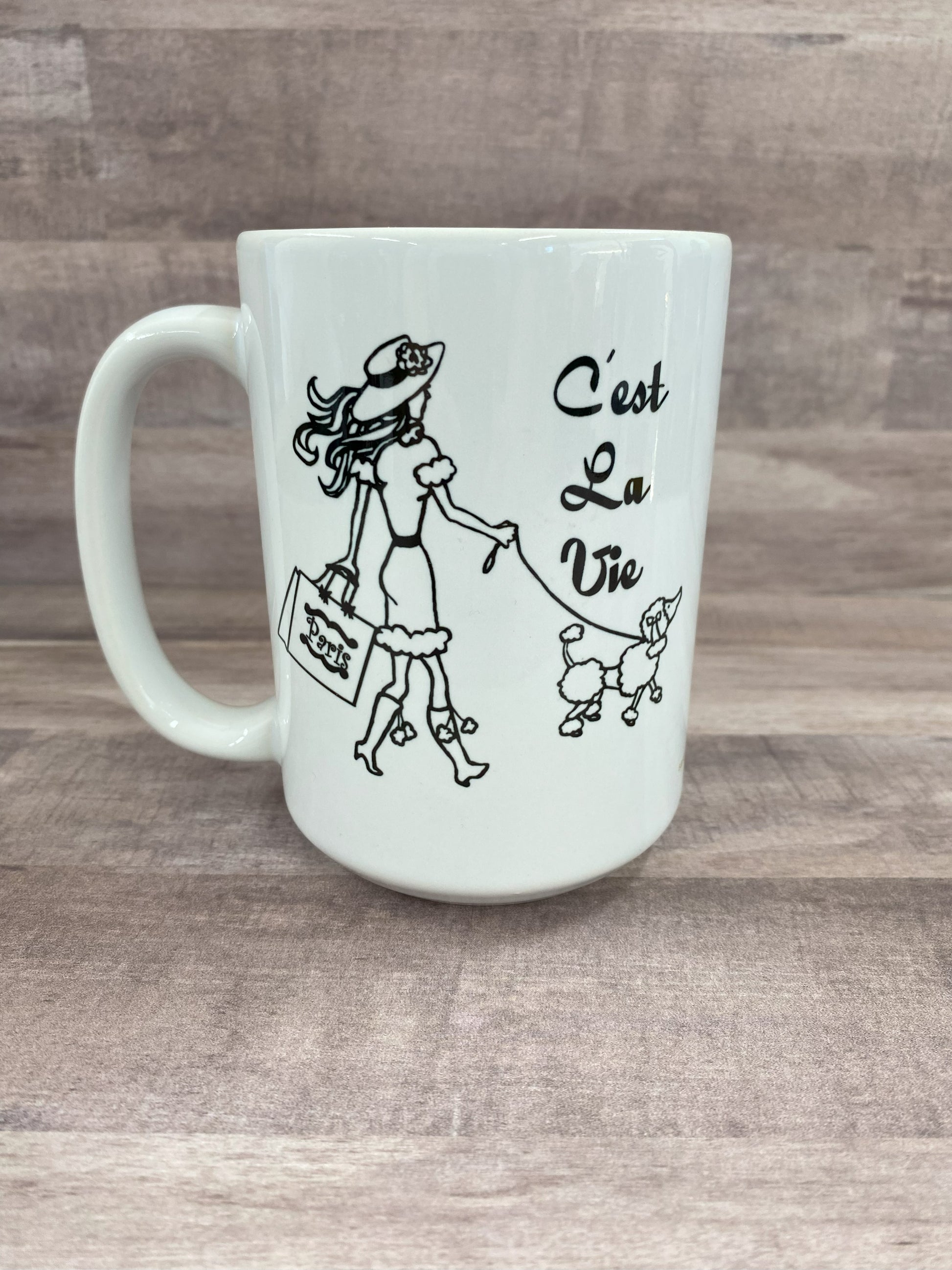 16oz cemaric white mug with the saying C'est la vie with an image of a woman carring shopping bags and walking a dog in blank sublimination ink.