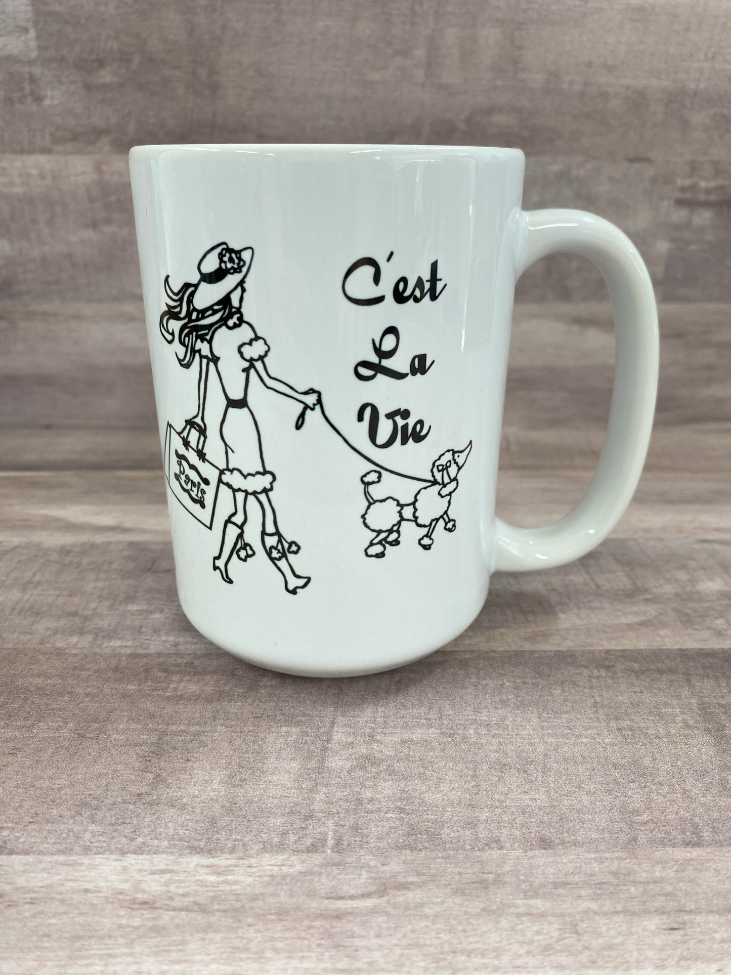 16oz cemaric white mug with the saying C'est la vie with an image of a woman carring shopping bags and walking a dog in blank sublimination ink.