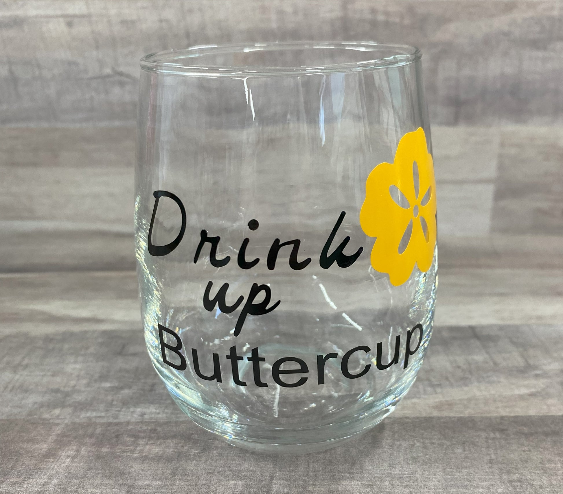 Stemless 15oz wine glass with the phrase drink up buttercup in black writing with a yellow flower. Customizable. Designed by Grit City Rebel.