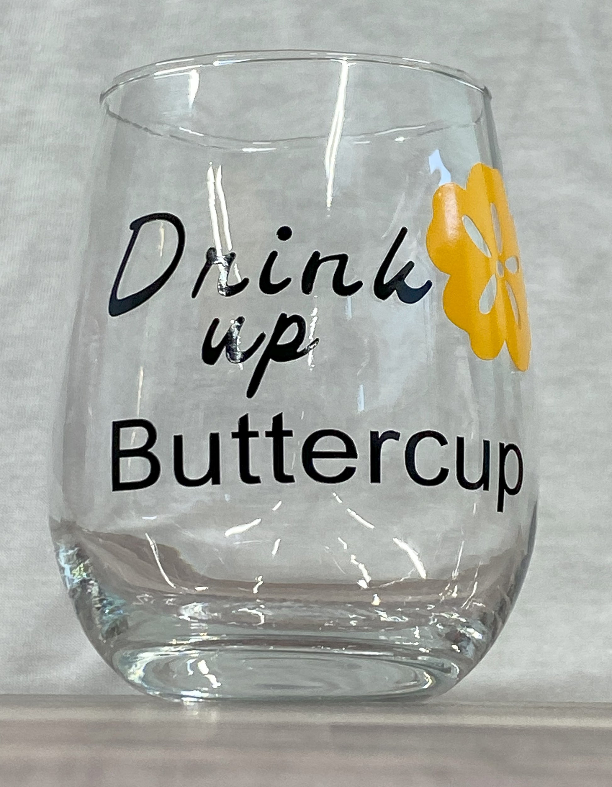 Stemless 15oz wine glass with the phrase drink up buttercup in black writing with a yellow flower. Customizable. Designed by Grit City Rebel.