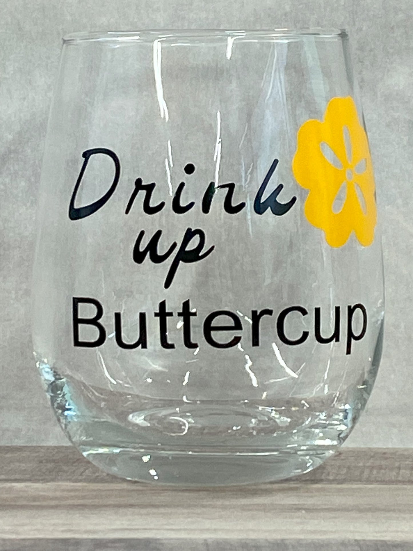 Stemless 15oz wine glass with the phrase drink up buttercup in black writing with a yellow flower. Customizable. Designed by Grit City Rebel.