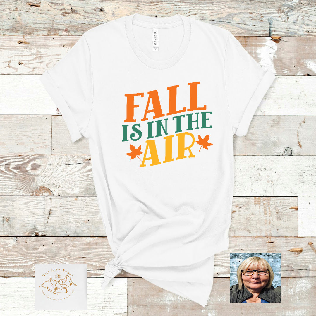 Fall is in the air saying in orange green and gols on a white Tshirt. Grit City Rebel sizing unisex sizes small medium large Xlarge 2X 3X