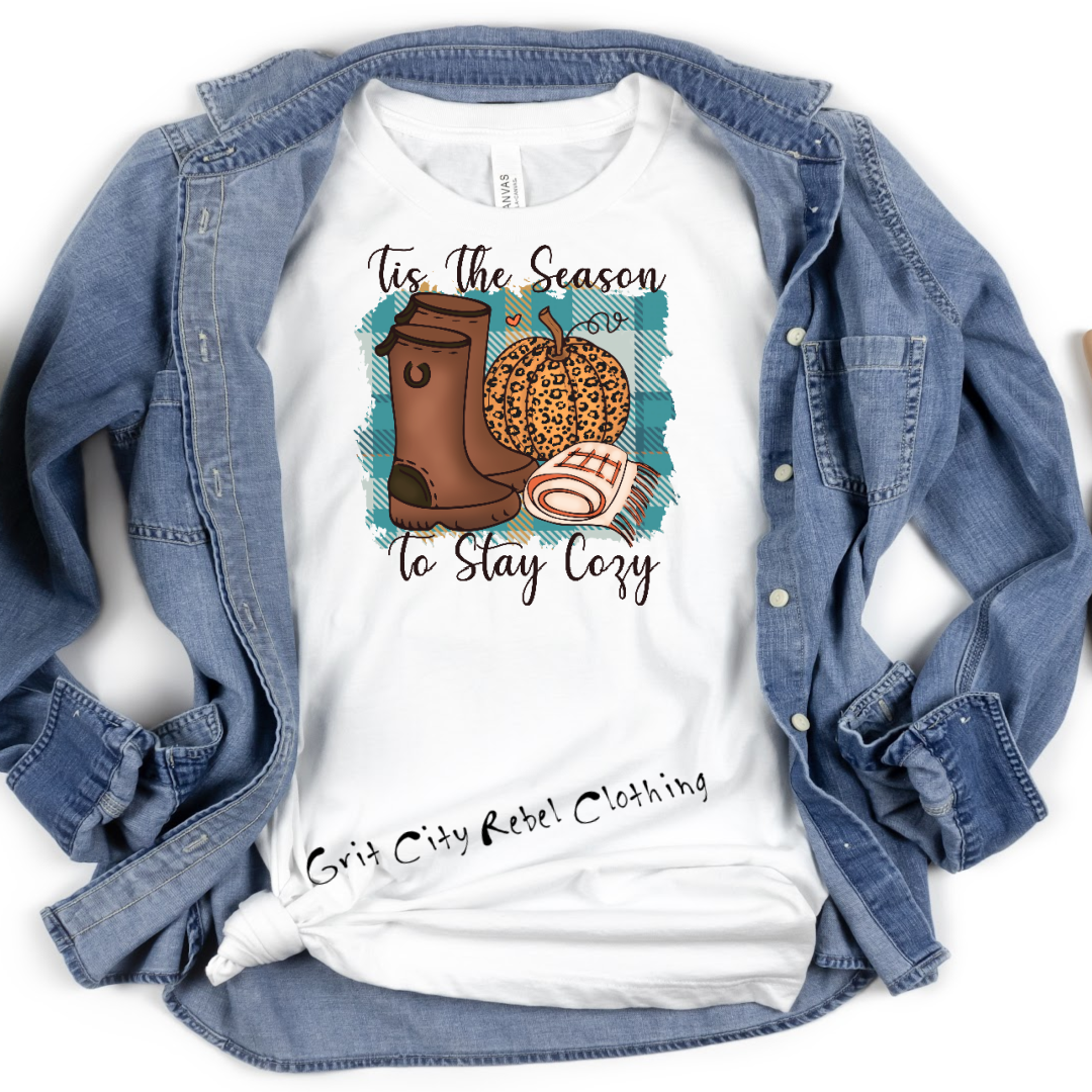 White Tshirt with teal and plaid background behind brown boots, leopard pumpkin and folded blanket, with the saying Tis The Season To Stay Cozy