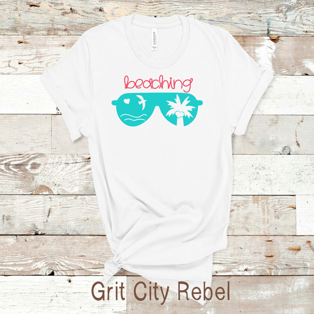 White short  sleeve T-Shirt with teal sunglasses that have a heart, palm tree, and bird, with the word beacjing in pink